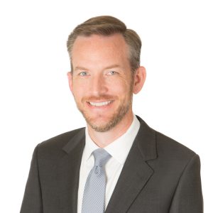 A smiling individual with a beard is wearing a gray suit, white shirt, and a tie against a plain background.