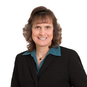 A smiling woman with shoulder-length hair wearing a black blazer over a teal blouse against a white background.
