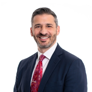 A smiling individual in professional attire featuring a dark blue suit and a red tie with a pattern.