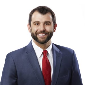 A smiling individual wearing a blue suit jacket, white shirt, and a red tie against a white background.
