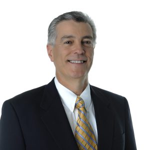 A person wearing a dark suit, a white shirt, and a yellow tie is smiling against a white background.
