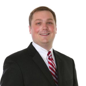 A smiling individual in professional attire with a black blazer and a striped red and white tie stands against a white background.