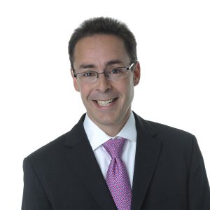 A smiling individual with glasses wearing a business suit and a pink patterned tie against a white background.