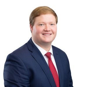 A person is wearing a dark suit with a red tie and a white shirt, smiling against a plain background.