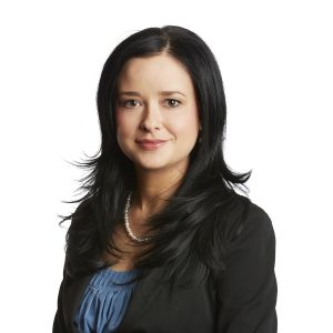 A professional-looking individual with dark hair is wearing a black blazer and a blue blouse, complemented by a pearl necklace.