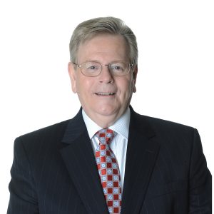A smiling individual wearing glasses, a dark suit, a white shirt, and a red tie with geometric patterns against a plain background.