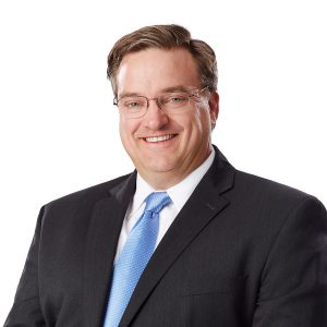 A smiling individual wearing glasses, a black suit, white shirt, and a light blue tie against a plain background.