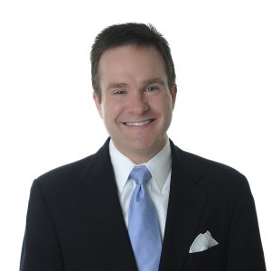 A smiling individual wearing a dark suit, light blue tie, and a white pocket square against a white background.