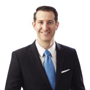 A smiling individual wearing a dark suit, light blue tie, and a white pocket square stands against a white background.