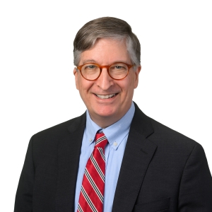 A man wearing a bowtie and glasses is smiling against a plain background.