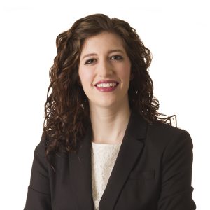 A smiling individual with curly hair wears a black blazer over a light-colored blouse against a white background.