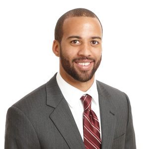 A smiling individual with a beard is wearing a gray suit and a red striped tie against a plain background.