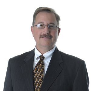 A man with a mustache is wearing glasses, a suit, and a patterned tie against a white background.