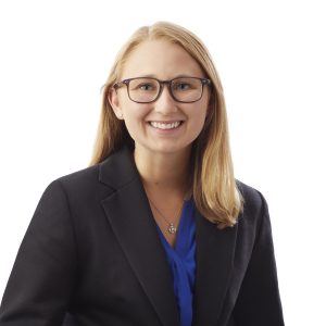 A smiling person with glasses wearing a dark blazer over a blue top against a white background.