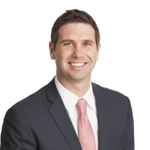 A smiling individual wearing a dark suit and a red patterned tie.