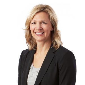 A smiling woman with shoulder-length blonde hair wearing a black blazer over a striped top against a white background.