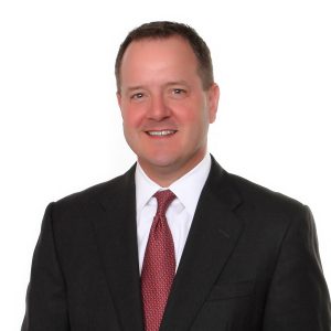 A man wearing a dark suit, white shirt, and red tie is smiling against a white background.