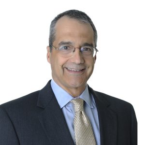 A smiling person wearing glasses, a dark suit, a light shirt, and a patterned tie poses for a professional portrait.