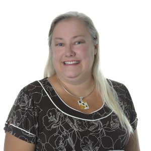 A smiling woman with long blonde hair wearing a black floral top and a necklace with an owl pendant.