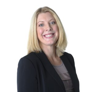 A smiling individual with blonde hair wearing a black blazer over a grey top against a white background.