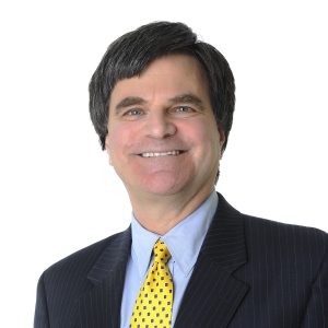 A smiling individual with dark hair wearing a dark pinstripe suit, white shirt, and yellow patterned tie against a light background.