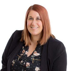 A smiling individual with reddish hair wearing a black blazer over a floral top is posing against a white background.