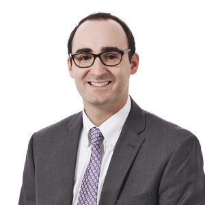 A smiling person wearing glasses, a gray suit, white shirt, and purple patterned tie.