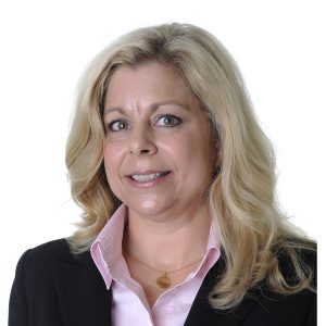 A person with blonde hair wearing a black blazer and a pink shirt against a white background.