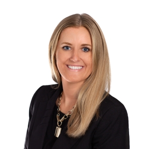 A smiling woman with long blonde hair wearing a black jacket and a necklace poses for a professional headshot.
