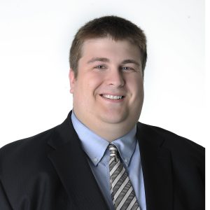 A smiling person dressed in a business suit with a striped tie against a plain white background.