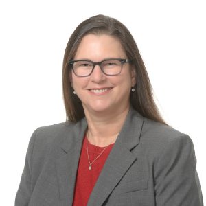 A smiling individual wearing glasses is dressed in business attire consisting of a gray blazer and a red shirt.