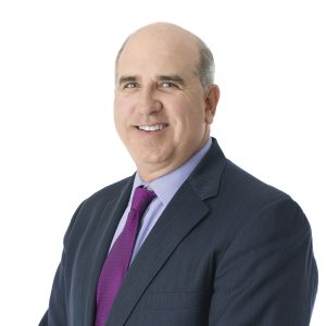 A balding individual is wearing a dark suit with a purple tie and smiling at the camera against a plain white background.