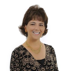 A smiling person with short hair wearing a patterned top and a gold necklace against a white background.
