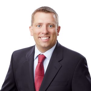 A smiling individual with short hair is wearing a dark suit, a checked shirt, and a red tie against a white background.
