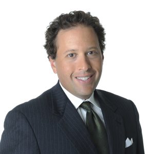 A man in a formal suit with a striped tie and pocket square is smiling at the camera.