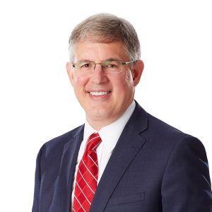 A smiling individual with glasses is wearing a navy blue suit and a red tie against a white background.