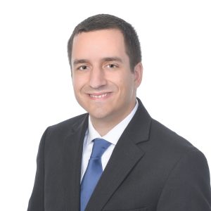 A smiling individual dressed in professional attire with a black blazer, white shirt, and blue tie poses against a plain background.