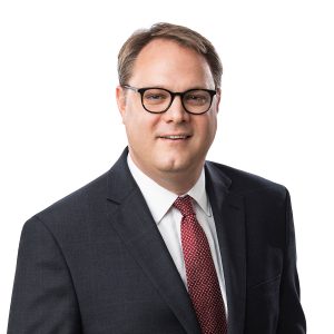 A smiling individual wearing glasses, a dark suit, white shirt, and a red patterned tie on a plain background.