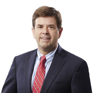 A person in a business suit with a red striped tie and a blue checked shirt against a white background.
