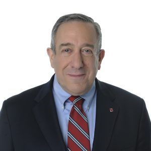 A person is wearing a suit with a red and grey striped tie, presenting a friendly smile, with a plain white background behind them.