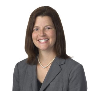 A smiling woman with shoulder-length brown hair wearing a gray suit jacket and a pearl necklace.