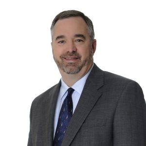 A person is wearing a grey suit, a white shirt, and a blue tie with a diagonal striped pattern, and is smiling at the camera against a plain background.