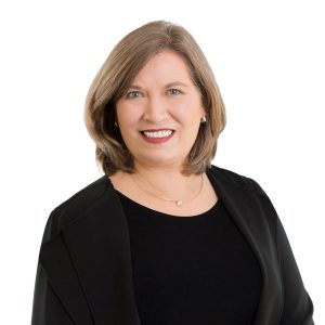 A smiling person with short hair is wearing a black outfit with a subtle necklace, against a white background.