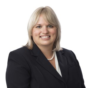 A smiling person with short blonde hair wearing a black blazer and a white top accessorized with a pearl necklace.