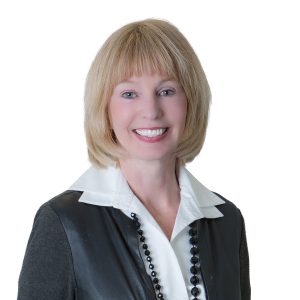 A smiling woman with blond hair wearing a black jacket, white shirt, and a pearl necklace against a white background.