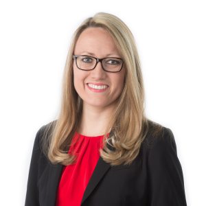 A smiling woman with blonde hair wearing glasses, a black blazer, and a red top stands against a white background.