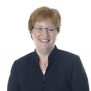 A smiling person with short hair is wearing a dark blazer over a light blouse against a white background.