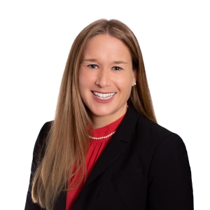 A smiling woman wearing a black blazer, red blouse, and a pearl necklace.