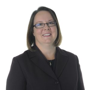 A smiling individual with glasses wearing a black business suit poses against a white background.