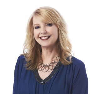 A smiling individual with blonde hair is wearing a blue top and a distinctive circular necklace.